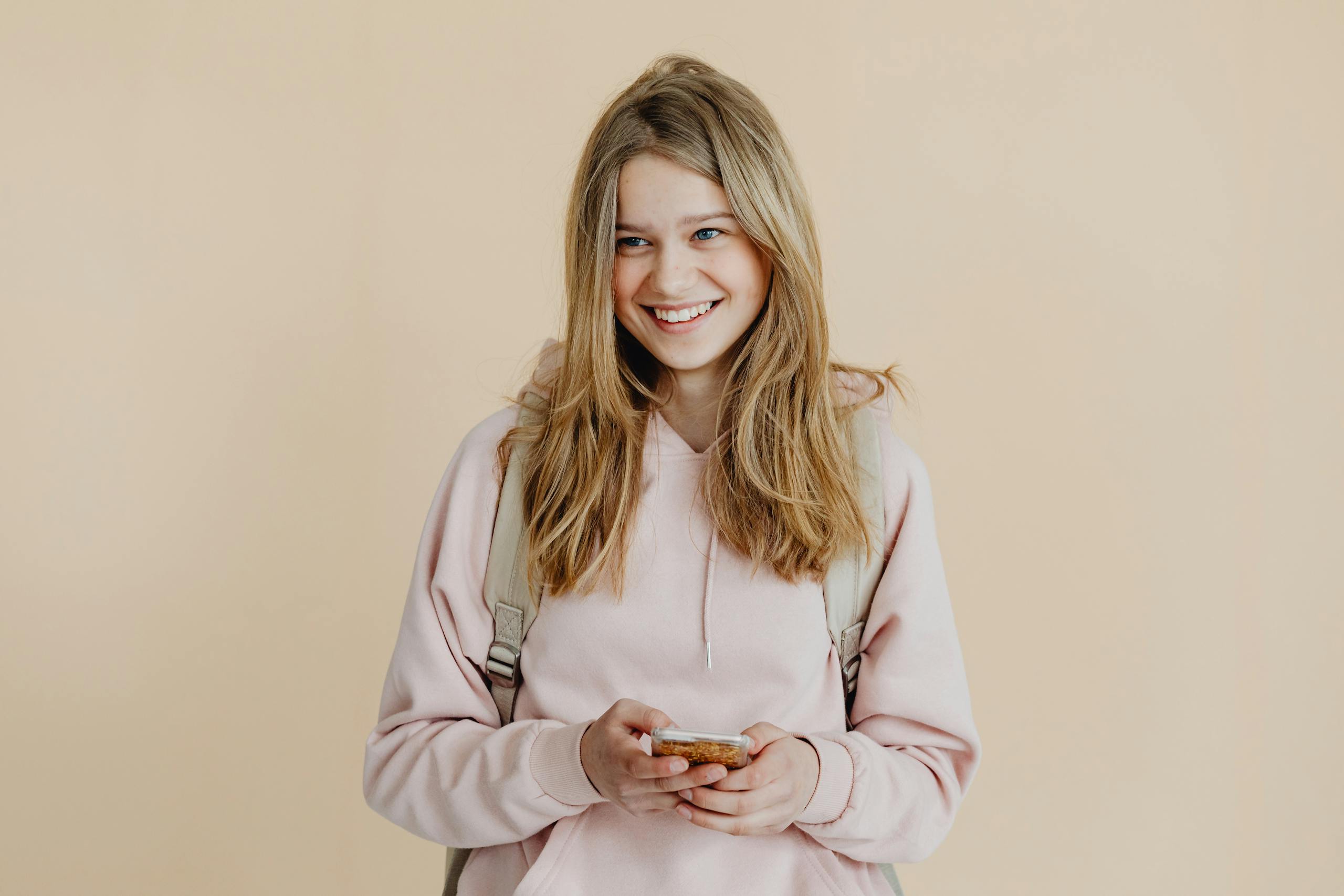A cheerful teenager with blonde hair wearing a pink sweater and using a smartphone indoors.