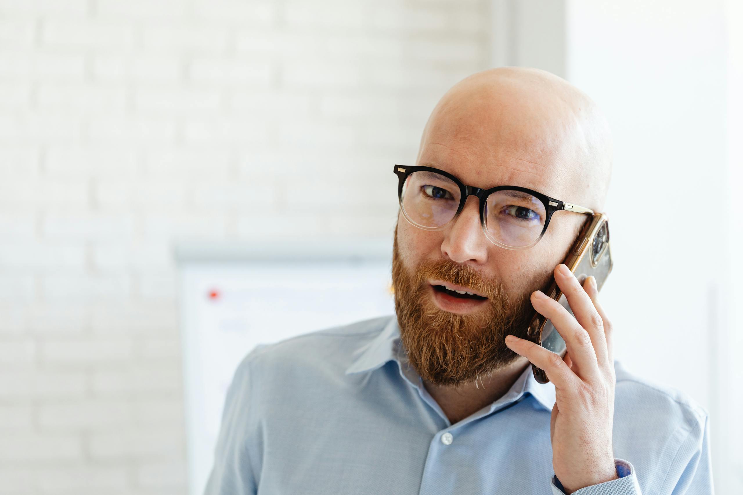 Bald bearded man in glasses using smartphone inside an office setting.